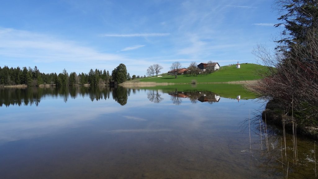 Bauernhof auf Hügel, davor ein See