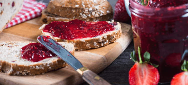 Frühstücksbrettchen mit zwei Marmeladebroten und Glas Marmelade, davor Erdbeeren