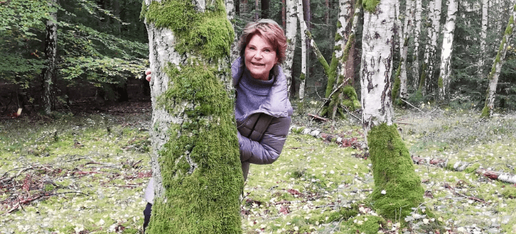 Vielleicht findet man den Sinn des Lebens im Wald. Eine Frau versteckt sich hinter einem Baum und lugt hervor.