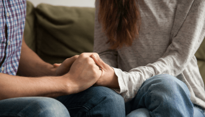 Ein Paar sitzt eng nebeneinander, nur Beine und Unterkörper sichtbar, sie halten sich an den Händen und zeigen gegenseitige Nähe und Zuwendung.